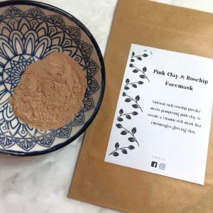 Bowl containing pink clay and rosehip powdered facemask next to facemask sachet