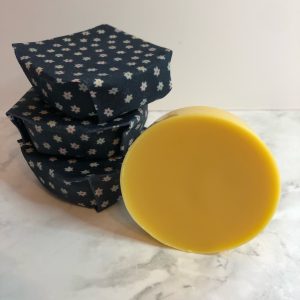 Stack of three solid hand cream bars wrapped in blue cotton beeswax wrap with small white flower imprint. Single, round bar of yellow hand cream stood on its side.